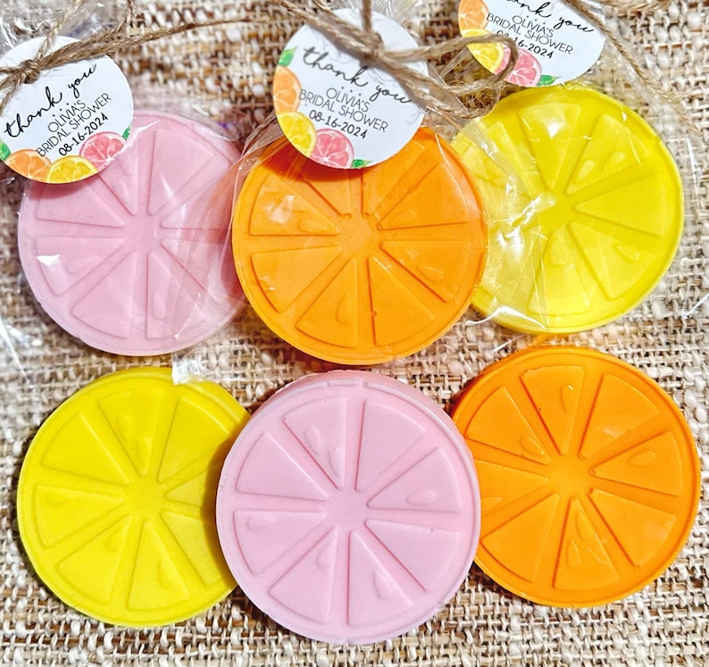 May include: Six round, citrus-shaped soaps in pink, yellow, and orange. The soaps are wrapped in clear plastic bags with thank you tags that say "Olivia's Bridal Shower" and the date.