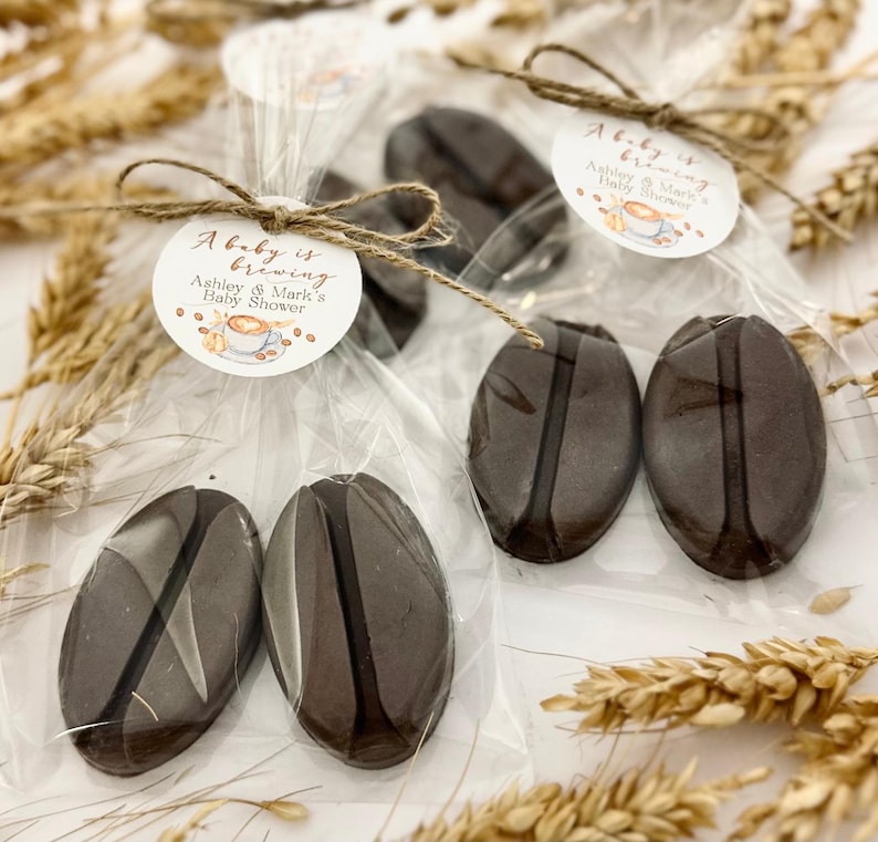 May include: Several cellophane-wrapped coffee bean-shaped favors tied with twine. Each favor has a circular tag that reads "A baby is brewing" and "Ashley & Mark's Baby Shower" with a coffee cup illustration. The favors are brown.