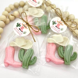 Op de afbeelding: Een set van drie kleine, roze en groene cadeautjes in een doorzichtige cellofaan zak, vastgebonden met touw. De cadeautjes zijn gevormd als een cowboylaars, een cactus en een cowboyhoed. Een rond label met de tekst "Thank y'all for coming!" is aan de zak bevestigd.