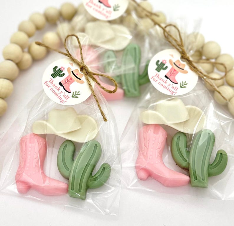 Op de afbeelding: Een set van drie kleine, roze en groene cadeautjes in een doorzichtige cellofaan zak, vastgebonden met touw. De cadeautjes zijn gevormd als een cowboylaars, een cactus en een cowboyhoed. Een rond label met de tekst "Thank y'all for coming!" is aan de zak bevestigd.