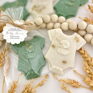 May include: Baby shower favors featuring onesie-shaped cookies in green and white, decorated with a duck design. One cookie is packaged in a clear bag with a tag that reads "Thank you for showering Baby Jones with love - Amy & John."