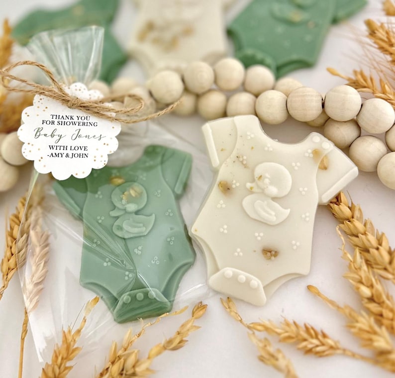 May include: Baby shower favors featuring onesie-shaped cookies in green and white, decorated with a duck design. One cookie is packaged in a clear bag with a tag that reads "Thank you for showering Baby Jones with love - Amy & John."
