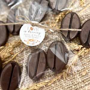 May include: A small clear bag filled with brown chocolate coffee bean shaped favors tied with twine and a round tag that says "Love is brewing Ashley & Mark's Bridal Shower".