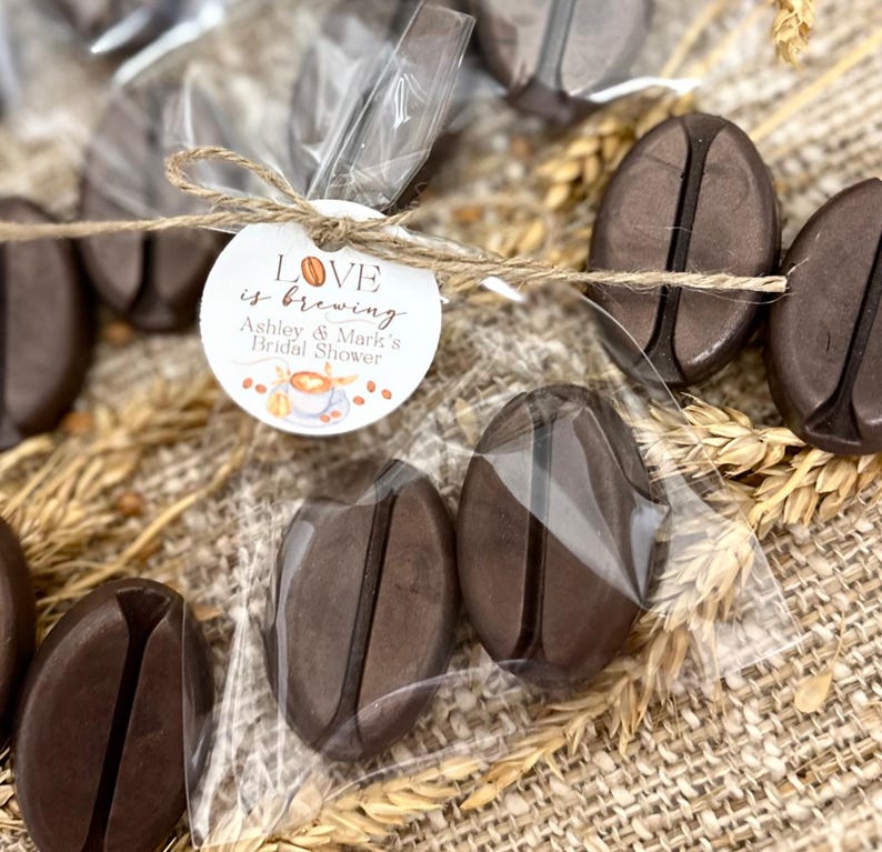 May include: A small clear bag filled with brown chocolate coffee bean shaped favors tied with twine and a round tag that says "Love is brewing Ashley & Mark's Bridal Shower".