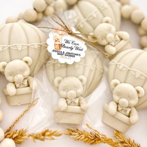 May include: A set of beige and white hot air balloon favors with teddy bears inside. The favors are tied with twine and have a tag that reads "We Can Bearly Wait Jessica & Jonathan's Baby Shower 08.16.25".