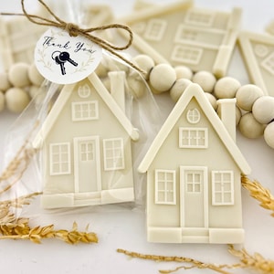 May include: Two off-white house-shaped favors in clear cellophane bags, tied with twine and a tag that reads "Thank You" with key illustrations. The houses have detailed windows, doors, and chimneys. Decorative wooden beads and wheat stalks are in the background.