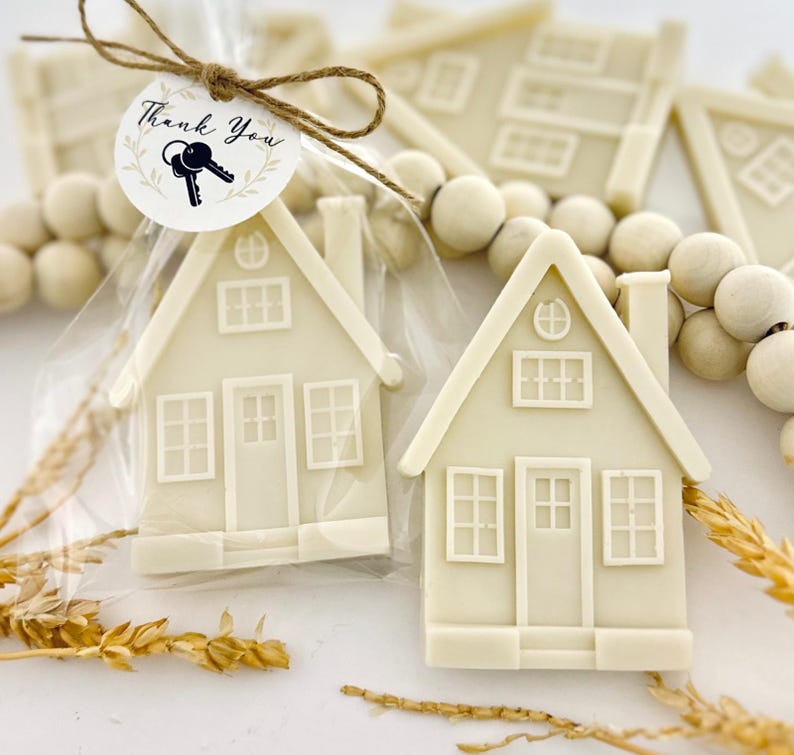 May include: Two off-white house-shaped favors in clear cellophane bags, tied with twine and a tag that reads "Thank You" with key illustrations. The houses have detailed windows, doors, and chimneys. Decorative wooden beads and wheat stalks are in the background.