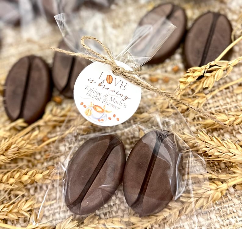 May include: Two brown coffee bean shaped soaps in a clear plastic bag tied with twine and a white circle tag that says "Love is brewing Ashley & Mark's Bridal Shower".