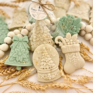 May include: A collection of molded wax shapes featuring a sage green bell with a poinsettia, a cream-colored snow globe with a tree, and a stocking with holly. A tag reads Happy Holidays FROM THE WALKERS 2026. Natural wheat stalks and wooden beads surround them.