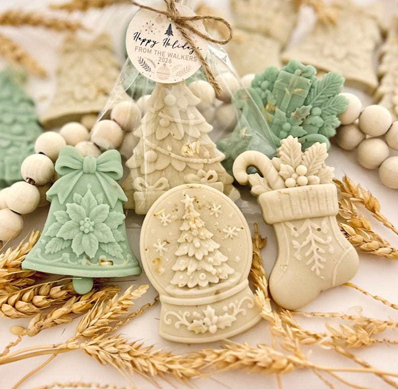 May include: A collection of molded wax shapes featuring a sage green bell with a poinsettia, a cream-colored snow globe with a tree, and a stocking with holly. A tag reads Happy Holidays FROM THE WALKERS 2026. Natural wheat stalks and wooden beads surround them.