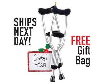 May include: A Christmas ornament shaped like a pair of crutches with a red and white sign that says "Cheryl Year". The ornament is silver and black with a red bow. The text "Ships Next Day!" and "Free Gift Bag" are also visible in the image.