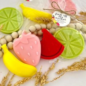 May include: Assortment of fruit-shaped soaps in a clear plastic bag with a "fresh off the market" tag. The soaps are yellow, pink, red, and green, and are shaped like a banana, strawberry, watermelon slice, and lime.