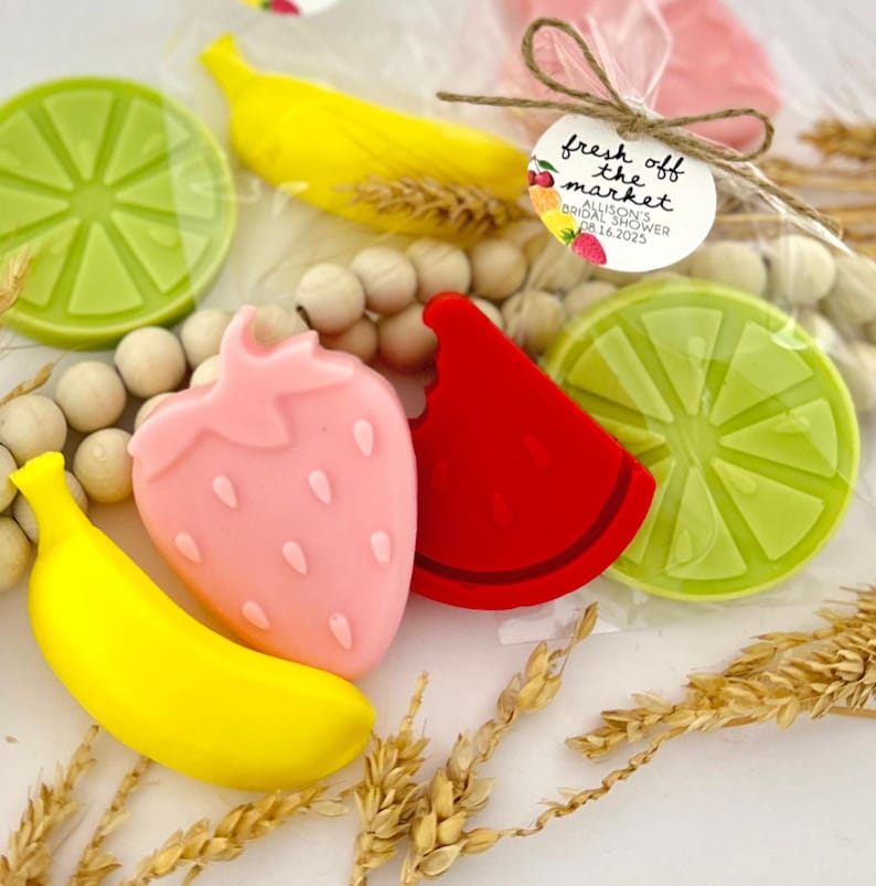 May include: Assortment of fruit-shaped soaps in a clear plastic bag with a "fresh off the market" tag. The soaps are yellow, pink, red, and green, and are shaped like a banana, strawberry, watermelon slice, and lime.