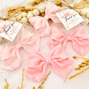 May include: Five pink fabric bows, each in a clear plastic bag, are arranged on a white surface. Each bow has a tag that reads "Thank you, Kayla's Baby Shower 08/12/21". Decorative elements include wooden beads and dried wheat.