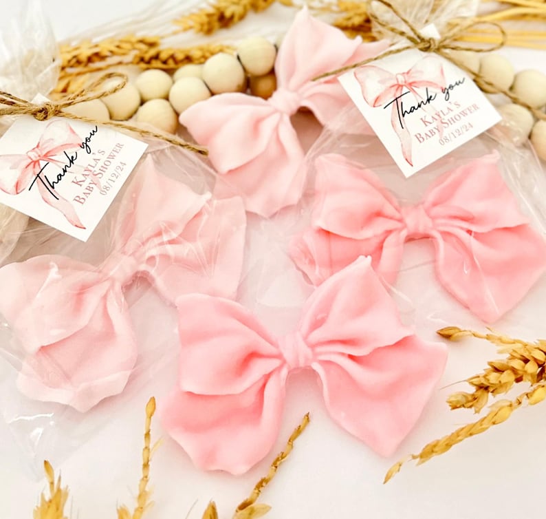 May include: Five pink fabric bows, each in a clear plastic bag, are arranged on a white surface. Each bow has a tag that reads "Thank you, Kayla's Baby Shower 08/12/21". Decorative elements include wooden beads and dried wheat.