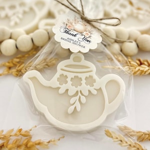 May include: A white teapot-shaped cookie with floral details, packaged in clear cellophane. A tag tied with twine reads "Thank You, Ashley's Bridal Shower" with floral accents. The background includes decorative beads and wheat stalks.