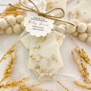 Op de afbeelding: Een baby shower gunst in de vorm van een romper, verpakt in doorzichtig cellofaan en vastgebonden met touw. De romper is wit met gouden accenten. Een label luidt "Bedankt voor het showeren van Baby Jones met liefde - Amy & John."