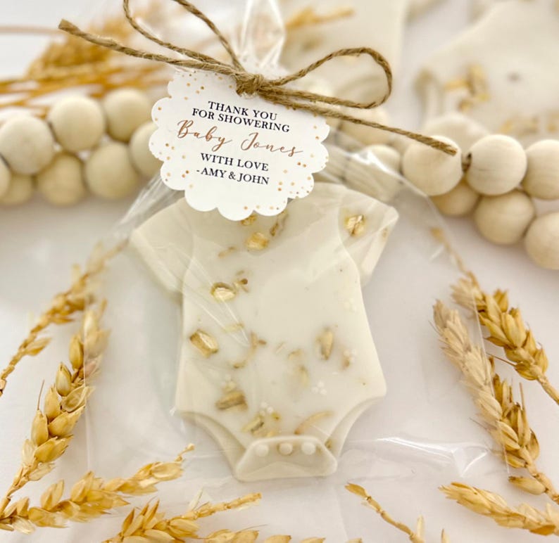 Op de afbeelding: Een baby shower gunst in de vorm van een romper, verpakt in doorzichtig cellofaan en vastgebonden met touw. De romper is wit met gouden accenten. Een label luidt "Bedankt voor het showeren van Baby Jones met liefde - Amy & John."