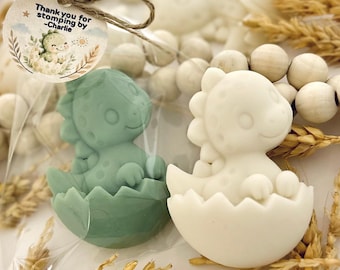 May include: Two dinosaur-shaped soaps, one sage green and one white, each emerging from a cracked egg. A round paper tag tied with twine reads, Thank you for stomping by -Charlie, featuring a small dinosaur illustration and wheat stalks.