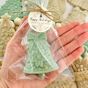 May include: A pale green bell-shaped wax melt inside a clear plastic bag, tied with twine and a round paper tag that reads Happy Holidays FROM THE WALKERS 2026. The wax features a molded bow at the top and a poinsettia flower design on the front.