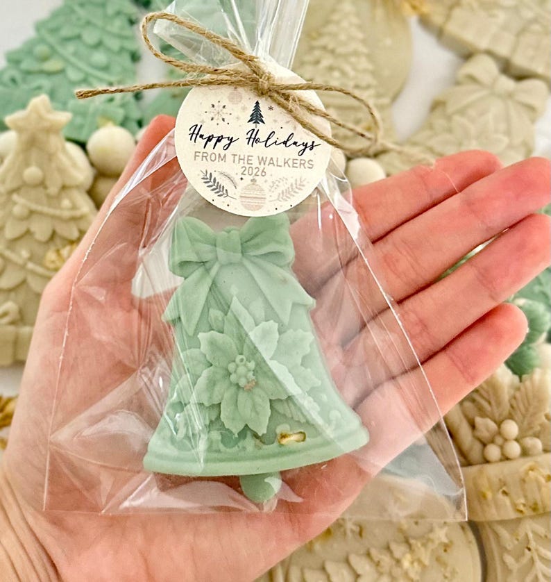 May include: A pale green bell-shaped wax melt inside a clear plastic bag, tied with twine and a round paper tag that reads Happy Holidays FROM THE WALKERS 2026. The wax features a molded bow at the top and a poinsettia flower design on the front.