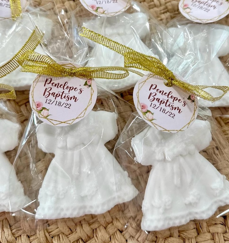 May include: White favour boxes with a white dress-shaped soap inside. Each box has a gold ribbon and a round tag with the text "Penelope's Baptism 12/18/22" in burgundy text with gold accents.