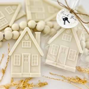 May include: Off-white house-shaped favors with detailed windows, doors, and chimneys. One favor is packaged in clear cellophane with a "Thank You" tag and key graphic. Wooden beads and wheat stalks are also visible.