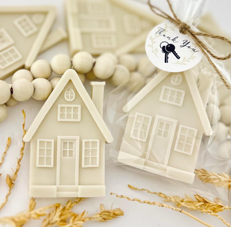 May include: Off-white house-shaped favors with detailed windows, doors, and chimneys. One favor is packaged in clear cellophane with a "Thank You" tag and key graphic. Wooden beads and wheat stalks are also visible.