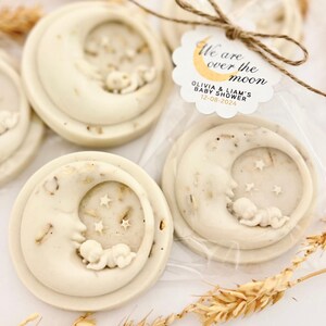 May include: White favor soaps shaped like a crescent moon with a baby sleeping inside. The soap is decorated with small white stars. The favor tag reads "We are over the moon Olivia & Liam's Baby Shower 12-08-2024"