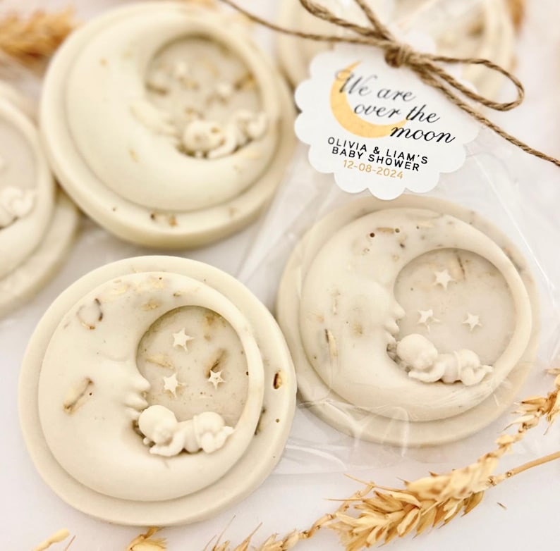 May include: White favor soaps shaped like a crescent moon with a baby sleeping inside. The soap is decorated with small white stars. The favor tag reads "We are over the moon Olivia & Liam's Baby Shower 12-08-2024"