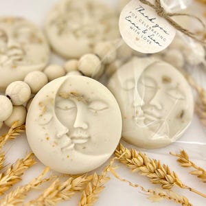 May include: Round, off-white soaps with a crescent moon and face design. Some soaps are packaged in clear cellophane with a small tag that reads "Thank you for celebrating Emma & Liam with Love." Wheat stalks and wooden beads are also present.