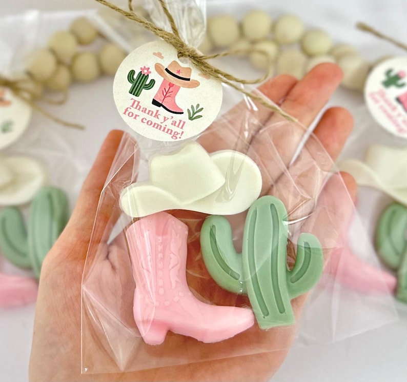 Op de afbeelding: Een doorzichtige plastic zak gevuld met kleine roze en groene zeepfavoren. De favoren zijn gevormd als cowboylaarzen, cactussen en cowboyhoeden. Een label dat aan de zak is bevestigd, luidt "Thank y'all for coming!"