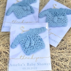 May include: A blue crocheted baby sweater on a hanger with a thank you tag that says "Amelia's Baby Shower 09.18.2022"