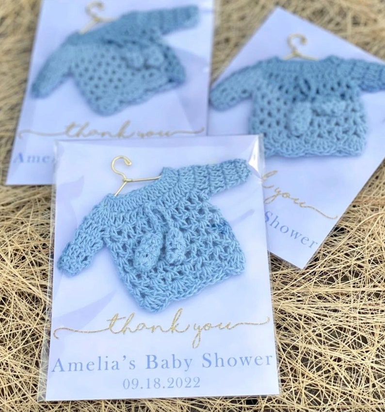 May include: A blue crocheted baby sweater on a hanger with a thank you tag that says "Amelia's Baby Shower 09.18.2022"