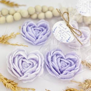 May include: Heart-shaped lavender-colored soaps in the shape of roses. The soaps are packaged in clear cellophane bags and tied with twine. A small round tag reads "Thank you for showering Katie with love!" The soaps are surrounded by decorative elements.