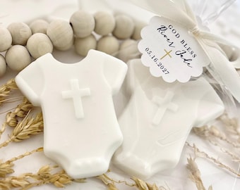 May include: Two white, baby-grow-shaped soaps featuring a raised cross design. One soap is wrapped in clear plastic with a white ribbon and a scalloped tag printed with "GOD BLESS River Jade 05.16.2027" and a small gold cross.