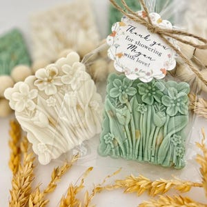 May include: Two floral-shaped soaps, one off-white and one green, are visible in clear cellophane. The soaps feature detailed flower and leaf designs. A tag tied with twine reads "Thank you for showering Megan with love!" Dried wheat stalks are in the foreground.