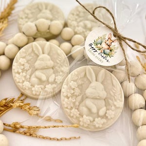 May include: Off-white Easter-themed soaps with a bunny and floral design. The soaps are round and packaged in clear cellophane. A round tag with a bunny and the text "Happy Easter From Avery" is tied to the package with twine. Decorative wooden beads and dried wheat stalks are also present.