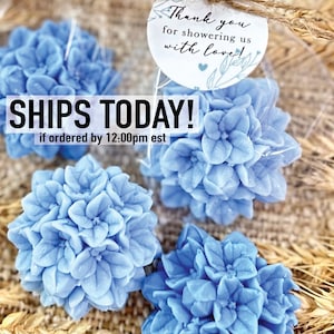 May include: Four blue hydrangea flower-shaped soaps wrapped in clear cellophane bags. The soaps are arranged on a woven brown background with wheat stalks. A white tag with the text "Thank you for showering us with love!" is attached to one of the bags.