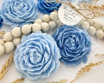May include: Light blue, rose-shaped soaps are the focal point of this image. Several soaps are visible, some individually wrapped. A tag tied with twine reads "Thank you for showering Olivia with love." Wooden beads and wheat stalks add to the presentation.