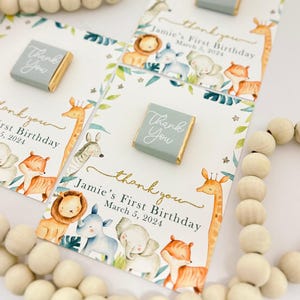 May include: Cards with watercolor illustrations of safari animals, including a giraffe, lion, and elephant. The cards say "Thank You" and "Jamie's First Birthday March 5, 2024". Small, light blue wrapped chocolates are also present.
