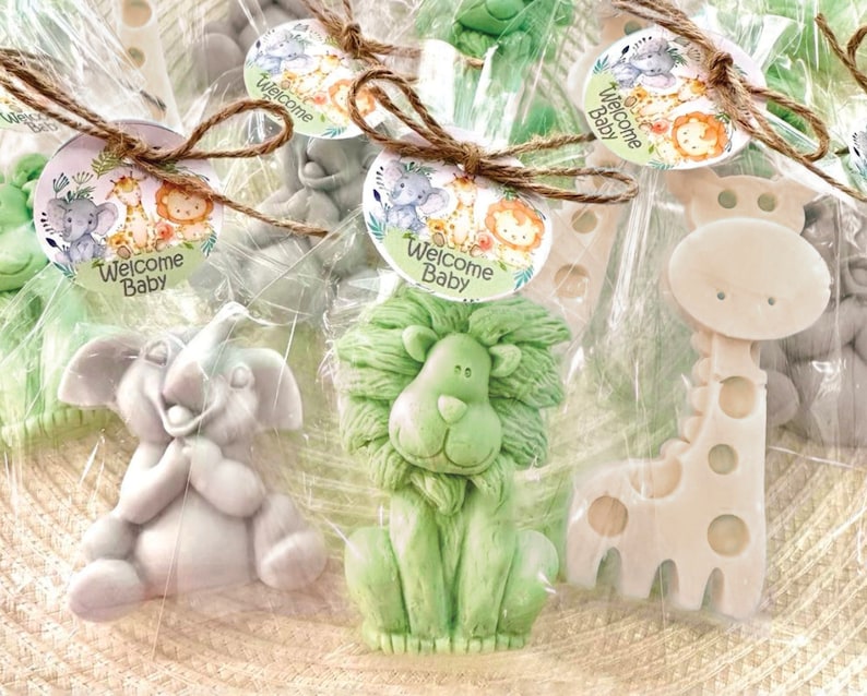 Puede incluir: Surtido de jabones peque&ntilde;os con forma de animales envueltos en bolsas de celof&aacute;n transparentes con un lazo de cuerda marr&oacute;n. Cada jab&oacute;n presenta un animal diferente, incluyendo un le&oacute;n, una jirafa y un elefante. Las bolsas est&aacute;n decoradas con etiquetas redondas y coloridas que dicen "Bienvenido Beb&eacute;".