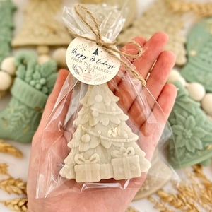 May include: A cream-colored Christmas tree-shaped wax melt inside a clear plastic bag tied with twine. A round paper tag is attached, featuring the text "Happy Holidays FROM THE WALKERS 2026" with small decorative illustrations of ornaments and foliage.