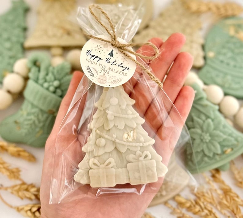 May include: A cream-colored Christmas tree-shaped wax melt inside a clear plastic bag tied with twine. A round paper tag is attached, featuring the text "Happy Holidays FROM THE WALKERS 2026" with small decorative illustrations of ornaments and foliage.