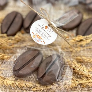 May include: Two brown coffee bean shaped favors in a clear plastic bag tied with twine and a white circle tag that says "LOVE is brewing Ashley & Mark's Bridal Shower".
