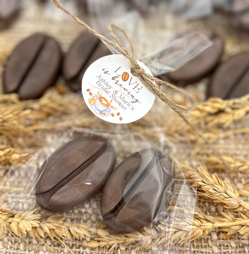 May include: Two brown coffee bean shaped favors in a clear plastic bag tied with twine and a white circle tag that says "LOVE is brewing Ashley & Mark's Bridal Shower".