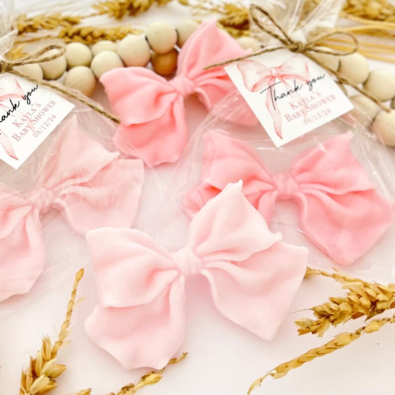 May include: Several light pink fabric bows, each tied with twine and packaged in clear plastic. Each bow has a small tag that reads "Thank you" and "Kayla's Baby Shower 08/12/24". The bows are arranged with decorative wooden beads and dried wheat.