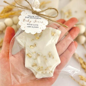 Op de afbeelding: Een baby shower gunst in de vorm van een witte romper, versierd met gouden accenten, wordt vastgehouden in een doorzichtige cellofaan zak. Een label vastgebonden met touw leest "THANK YOU FOR SHOWERING Baby Jones WITH LOVE -AMY & JOHN."