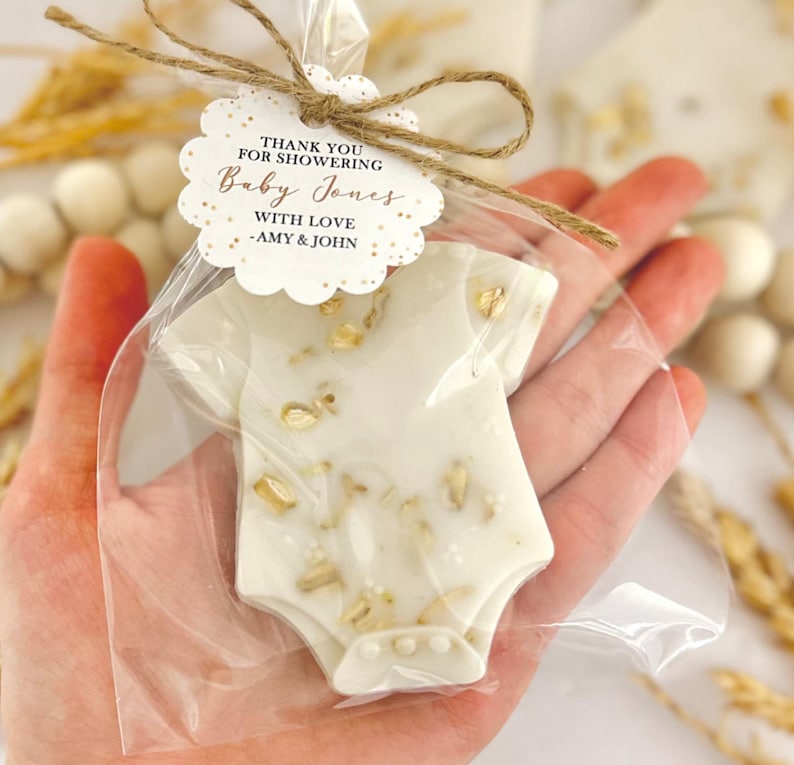 Op de afbeelding: Een baby shower gunst in de vorm van een witte romper, versierd met gouden accenten, wordt vastgehouden in een doorzichtige cellofaan zak. Een label vastgebonden met touw leest "THANK YOU FOR SHOWERING Baby Jones WITH LOVE -AMY & JOHN."