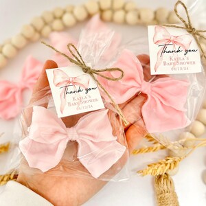 May include: Pink bow-shaped favors in clear cellophane bags, tied with twine and small tags. The tags read "Thank you, Kayla's Baby Shower 08/12/24". The bows are a soft, pastel pink color, perfect for a baby shower.